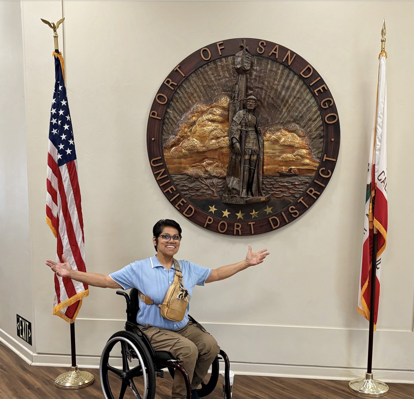 A person in a manual wheelchair poses cheerfully with open arms in front of the official round wooden seal of the Port of San Diego, inside the Port Administration Building, bordered by two standing flags.